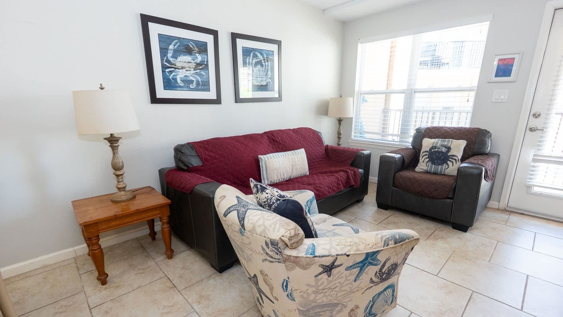 A living room with a red sofa, a patterned armchair, and a black armchair with a crab pillow. There are two framed artworks on the wall, a wooden side table with a lamp, and a window with blinds.