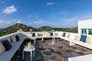 A rooftop terrace with a seating area, a table with drinks, and a view of a hilly landscape. - Casa Mare – Modern 5-Bedroom St. John Villa with Private Pool, Ocean Views & Rooftop Stargazing (cruz bay)