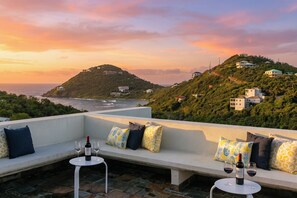 A rooftop terrace with a seating area, a table with wine glasses, and a scenic view of the ocean and hills. - Casa Mare – Modern 5-Bedroom St. John Villa with Private Pool, Ocean Views & Rooftop Stargazing (cruz bay)