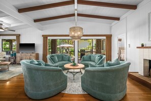 Ein geräumiges Wohnzimmer mit hoher Decke, einem großen Kronleuchter und einem Kamin. In der Mitte befinden sich zwei grüne Sessel und ein Couchtisch.