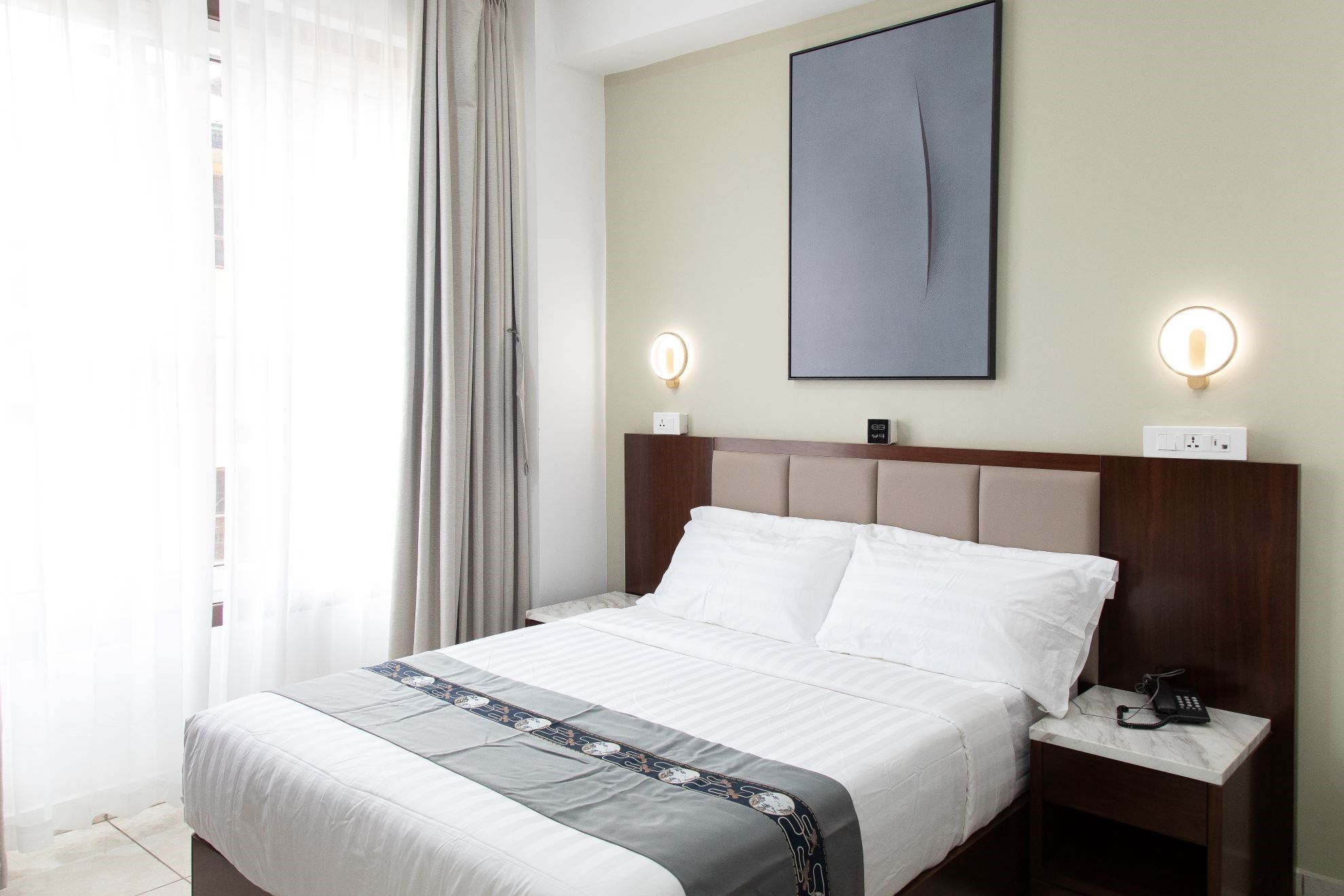 A hotel room with a bed, bedside tables, a wall-mounted light, and a painting.