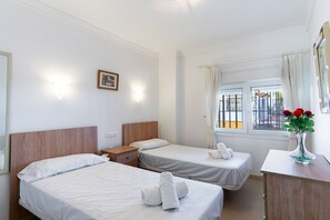A hotel room with two single beds, a wooden headboard, a nightstand, a small table, a vase with flowers, and a window with curtains. - Carabeo 10 by Casasol (Nerja)