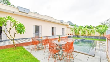 A poolside area with a table and chairs, a small pool, and a garden with a lawn and trees.