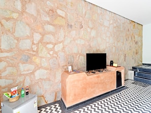 A room with a stone wall, a flat-screen TV on a wooden stand, a small refrigerator, and a patterned floor. - Dancenter Parijat 4bhk Pvt Villa I Pool I Udaipur (Udaipur)