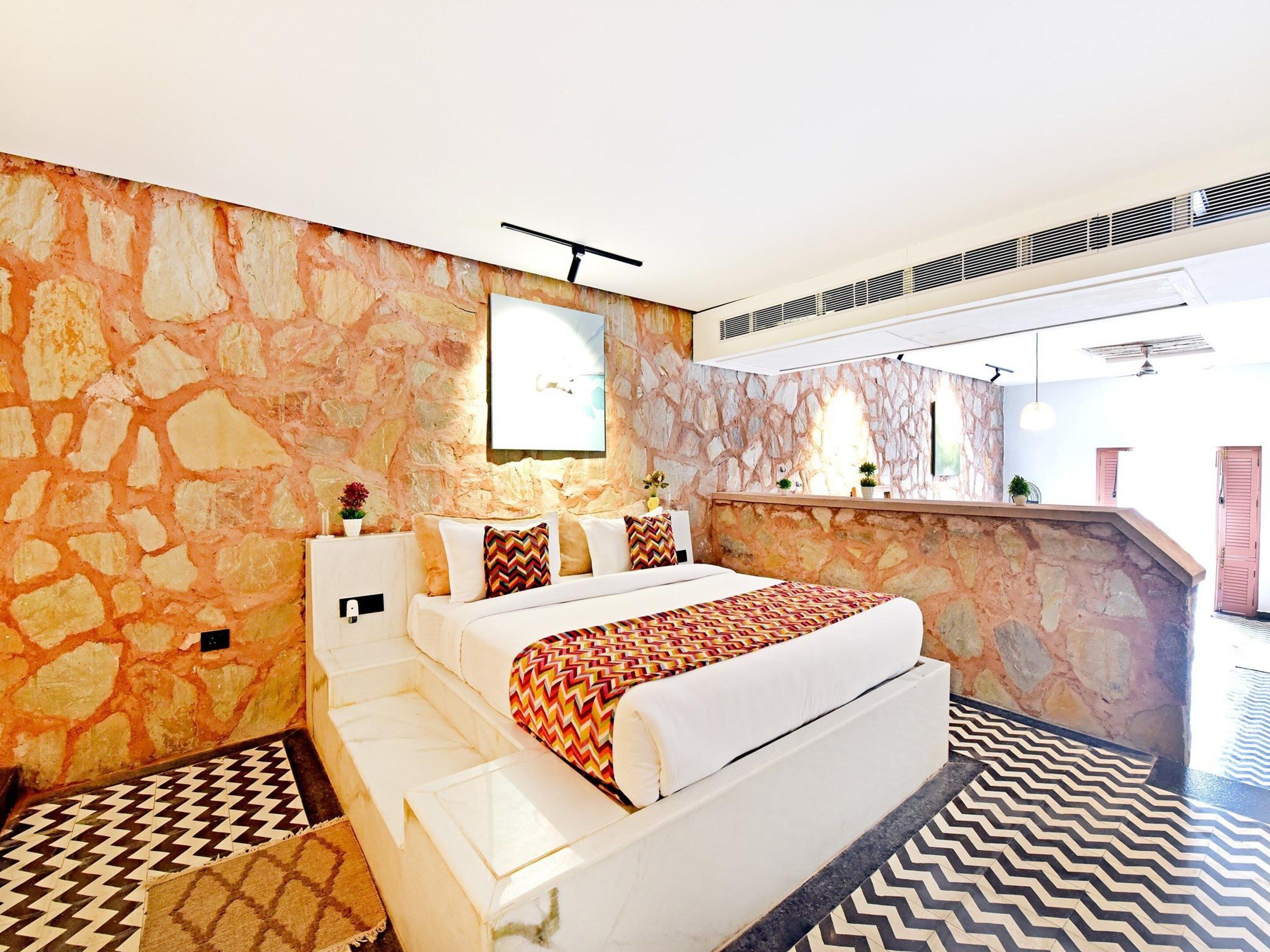 A hotel room with a stone wall, a bed with patterned bedding, a small table, and a view of the outside.