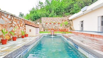 Una piscina con un muro de piedra y plantas en macetas en un lado, y un edificio con una puerta en el otro.