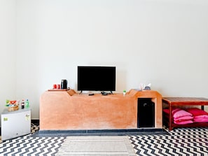 A room with a television on a stand, a small refrigerator, a bed with pink pillows, and a patterned rug. - Dancenter Parijat 4bhk Pvt Villa I Pool I Udaipur (Udaipur)
