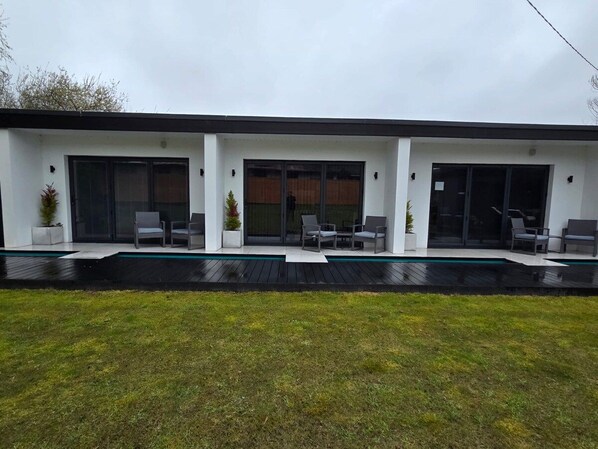A modern two-story house with a flat roof, large sliding glass doors, and a covered outdoor seating area with chairs. - Luxury 3-bed home with hot tub, full size snooker table & outdoor kitchen (Goonhavern)