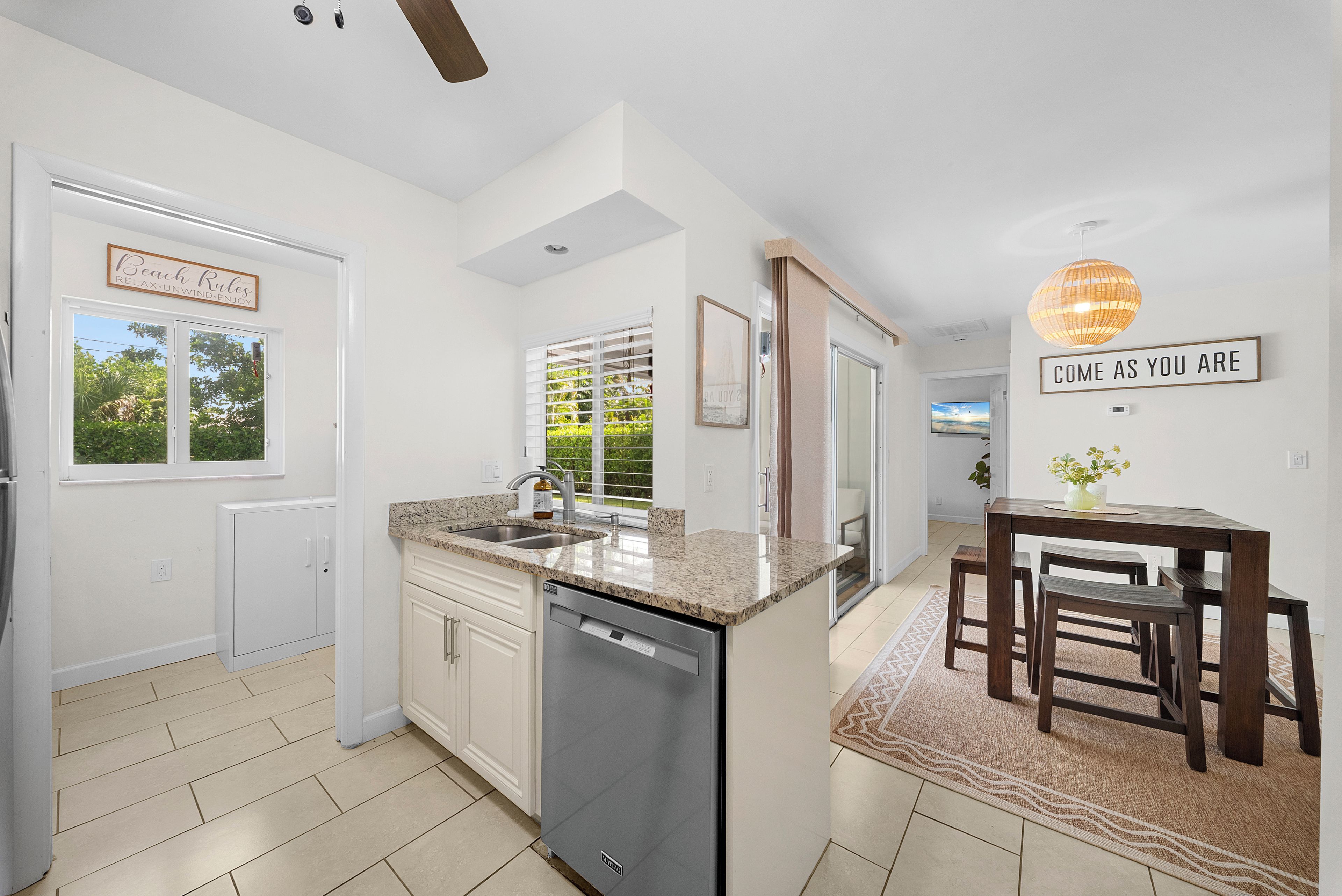 A modern kitchen with a dishwasher, a dining area with a table and chairs, and a view of greenery outside.