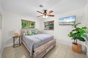 A bedroom with a wooden bed, a ceiling fan, a window with blinds, a bedside table with a lamp, and a wall-mounted TV. - 2BR Queen Suites • Heated Pool & BBQ • Minutes to the Beach (Naples)