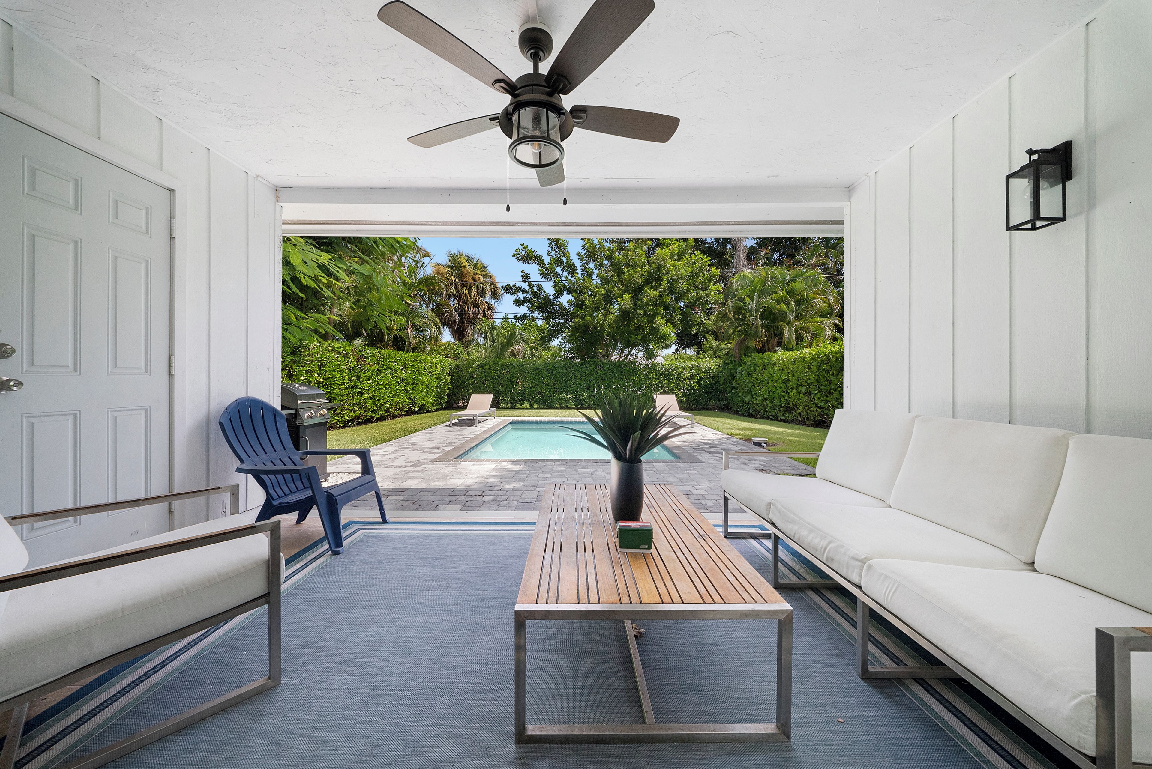 A covered patio area with a ceiling fan, a pool, a white sofa, a blue Adirondack chair, and a wooden table.