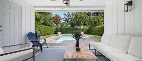 A covered patio area with a ceiling fan, a pool, a white sofa, a blue Adirondack chair, and a wooden table.