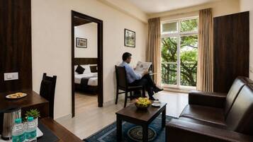 Habitación de hotel con cama, zona de estar con mesa y sillas, ventana con vista a árboles y puerta que da a otra habitación.