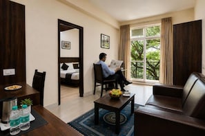 A hotel room with a bed, a sitting area with a table and chairs, a window with a view of trees, and a door leading to another room. - AVM COMFORT INN (Bengaluru)