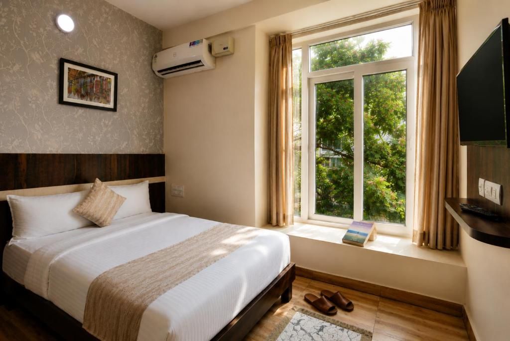 Ein Hotelzimmer mit Bett, einem Fenster mit Vorhängen, einer Klimaanlage, einem gerahmten Bild an der Wand und einem Fernseher auf einem Standfuß.