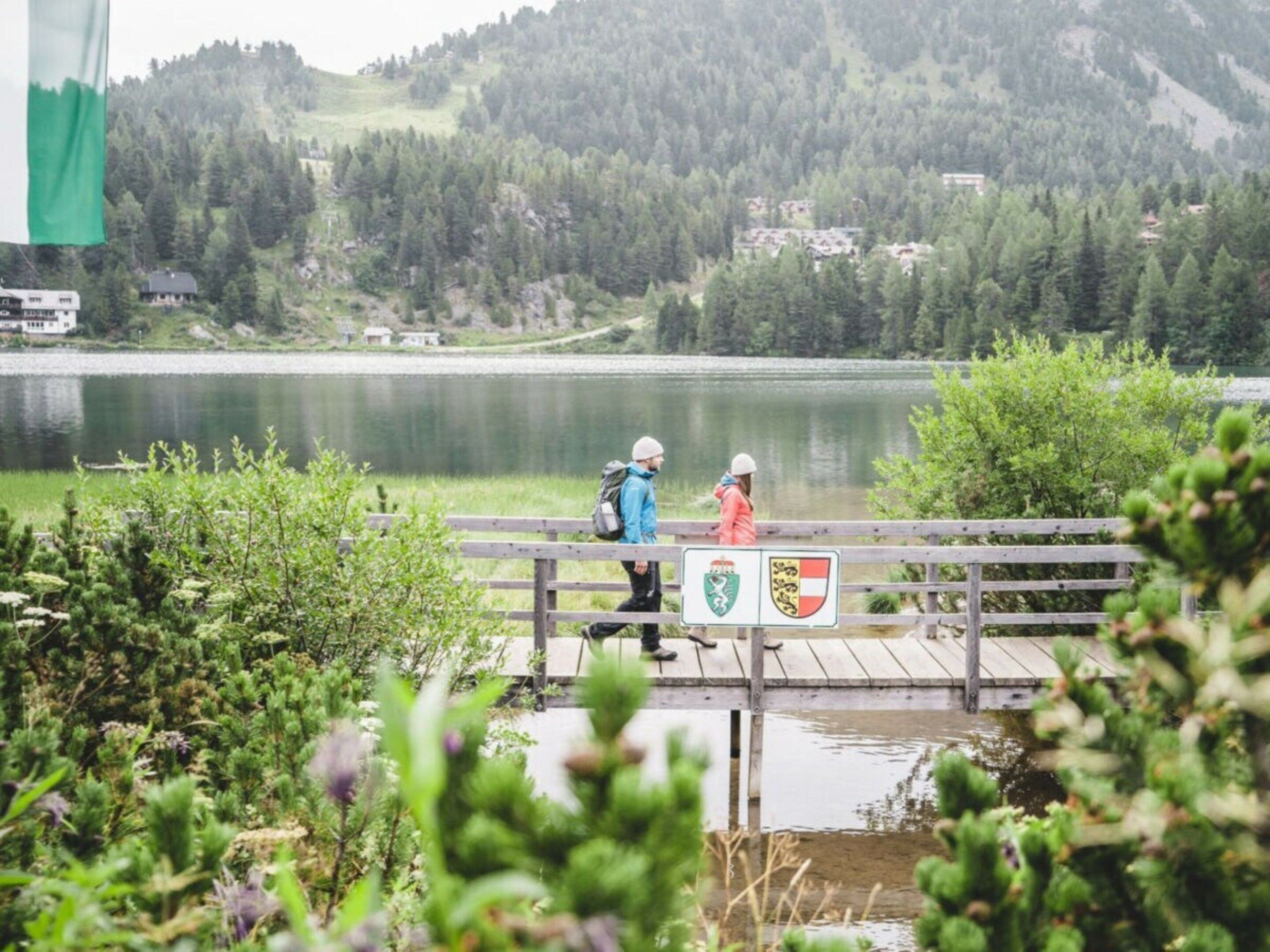 Eine Holzbrücke über einem See, auf der sich zwei Personen befinden, umgeben von viel Grün und einer Berglandschaft.