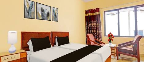 A hotel room with a double bed, a bedside table with a lamp, a chair, a small table with a vase, and a window with curtains.