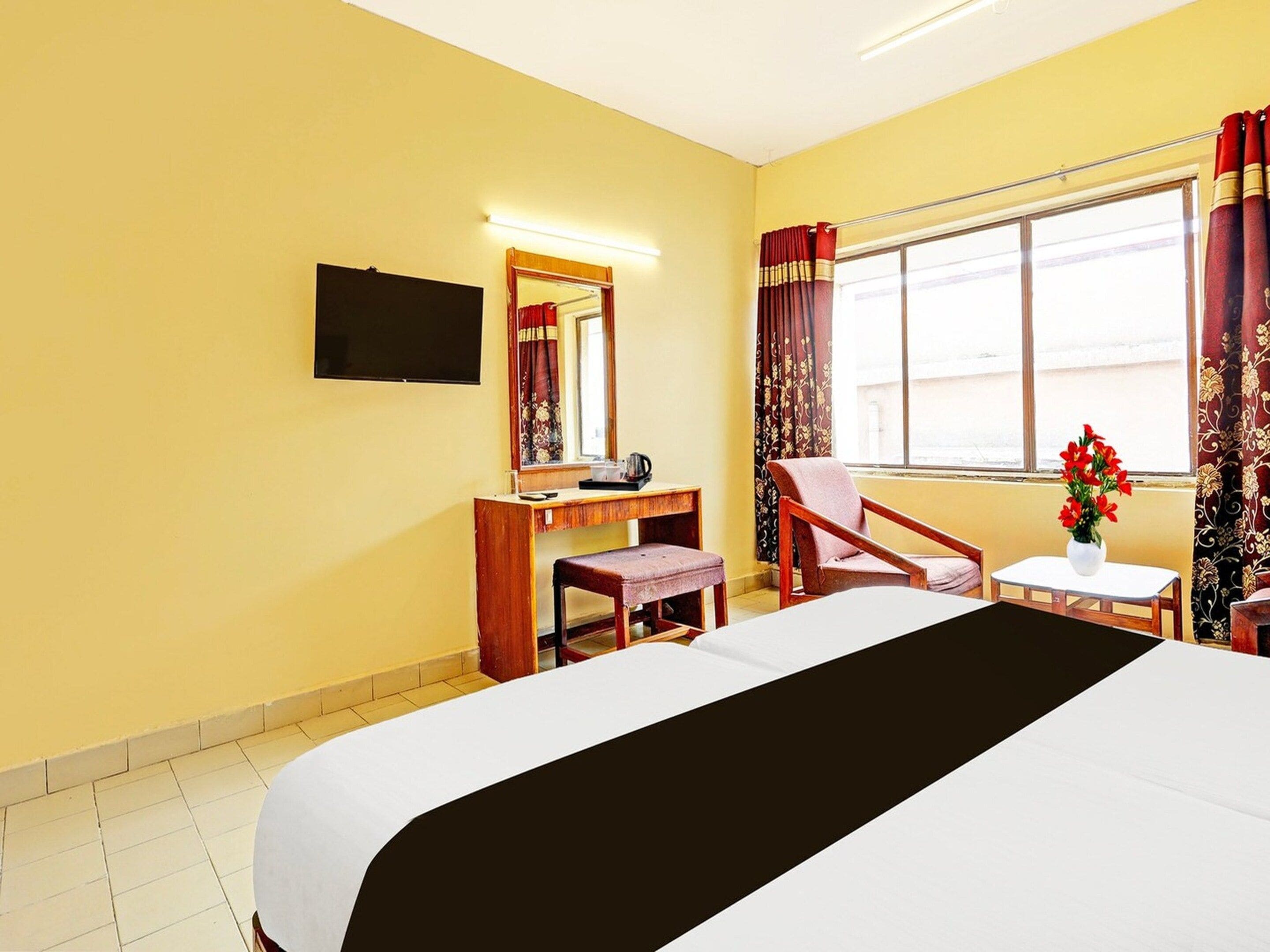 A hotel room with a bed, a desk, a chair, a TV, a window with curtains, and a vase with flowers.