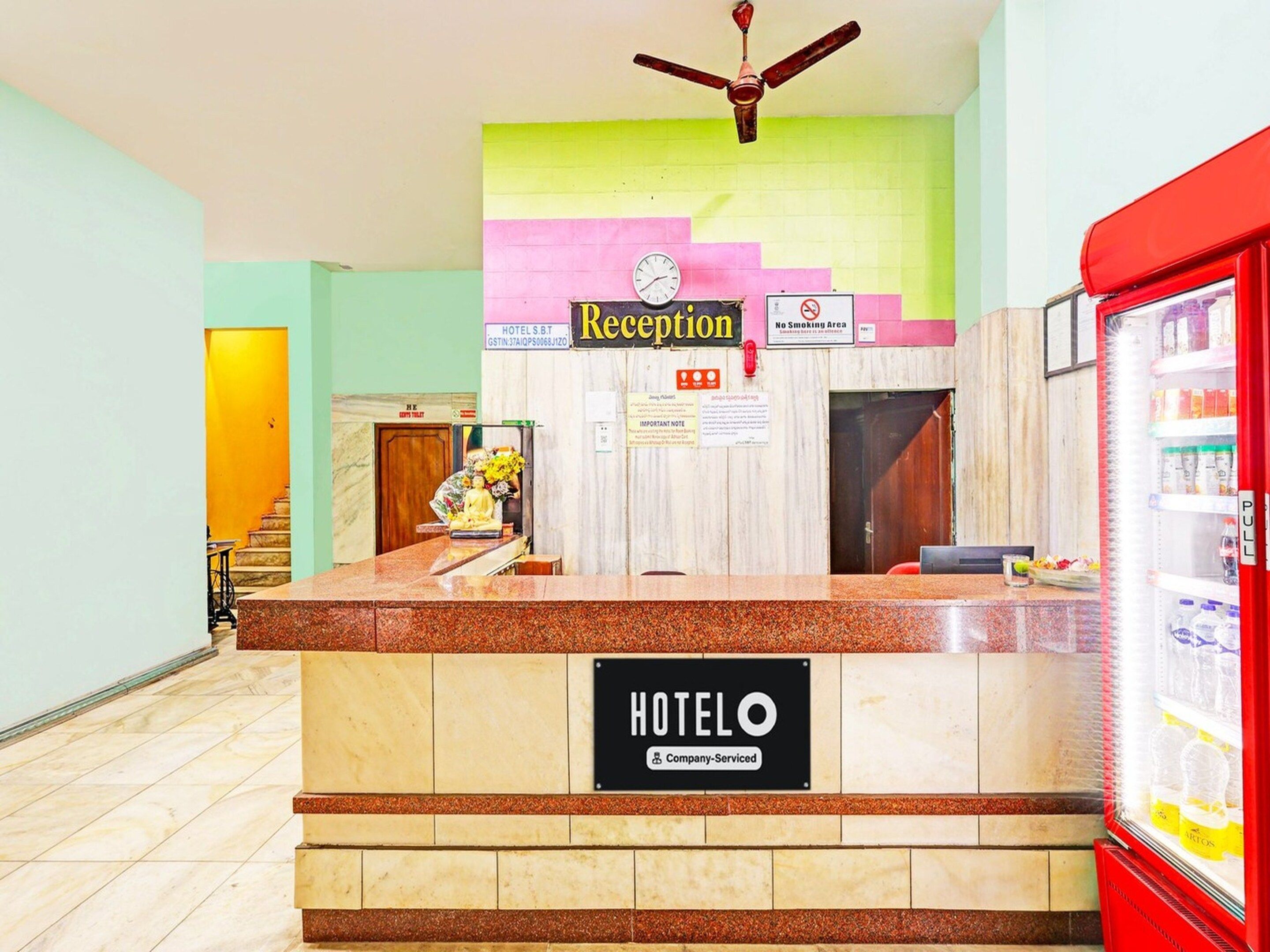 Un mostrador de recepción de hotel con un cartel de recepción, un reloj, un letrero de no fumar y un refrigerador con bebidas.