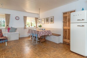 Ett kök med ett vitt kylskåp, träskåp, ett matbord med stolar, en bänk och ett fönster med gardiner.
