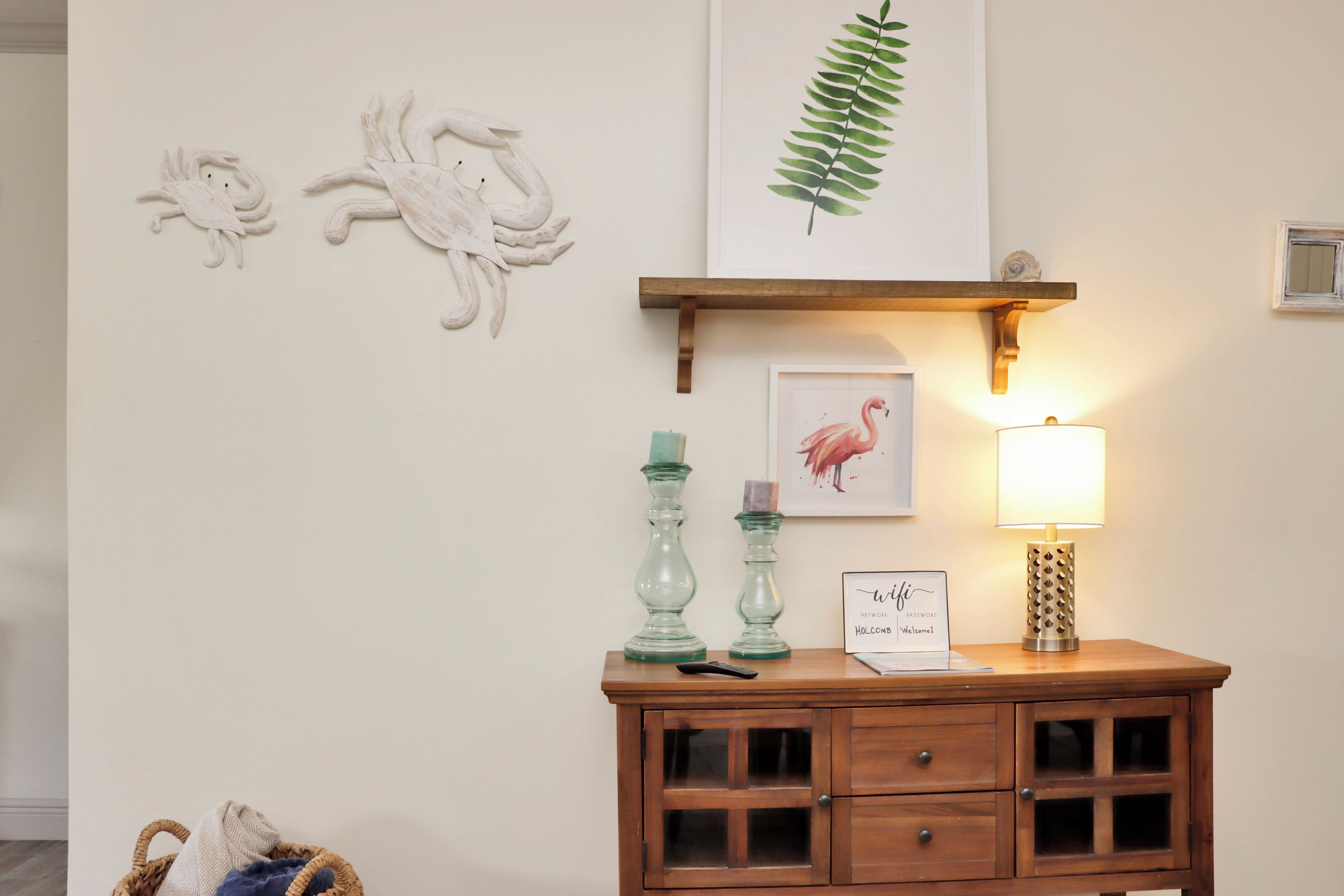 A wooden cabinet with glass doors, a framed welcome sign, a wall-mounted shelf with a framed leaf, and wall art of a flamingo and a bird.