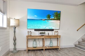 A living room with a large flat-screen TV mounted on the wall, a wooden console table with decorative items, a lamp, and a staircase. - Steps to Beach | Great Location | Fenced in Yard | Mystic Nights by the Sea (Mexico Beach)