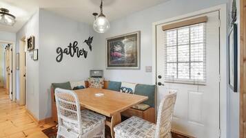 Un'area pranzo con un tavolo in legno e sedie in vimini, una parete con il cartello 'gather', un quadro incorniciato e una finestra con persiane.