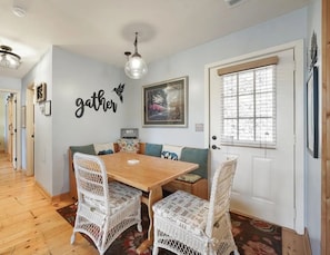 Un'area pranzo con un tavolo in legno e sedie in vimini, una parete con il cartello 'gather', un quadro incorniciato e una finestra con persiane.