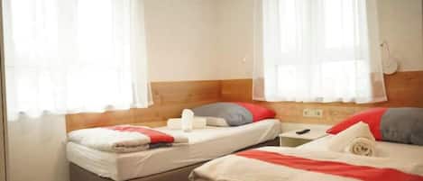 A room with two beds, each with a pillow and a blanket. There are curtains on the windows, and a lamp on the wall.