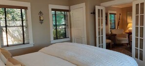 Un dormitorio con una cama grande, dos ventanas con persianas, una puerta que da a otra habitación y una lámpara en una mesita de noche.