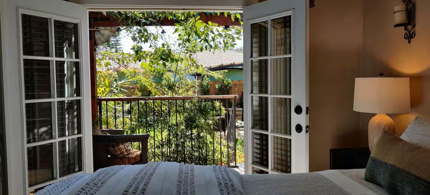 Une chambre à coucher avec un lit, une table de chevet, une lampe et une vue sur un jardin, grâce à des portes ouvertes.