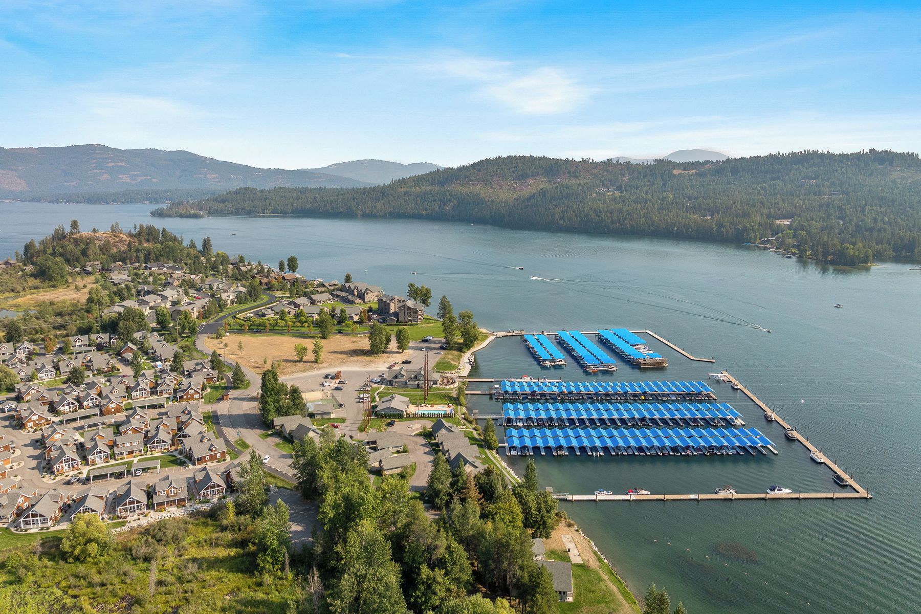 An aerial view of a lakeside community with residential housing, a marina with docked boats, and a forested backdrop.