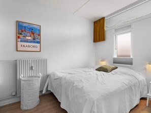 A minimalist bedroom with a bed, a radiator, a window with curtains, a wall-mounted light, and a framed picture of a town. - 4 Star Holiday Home in Faaborg (Faaborg)