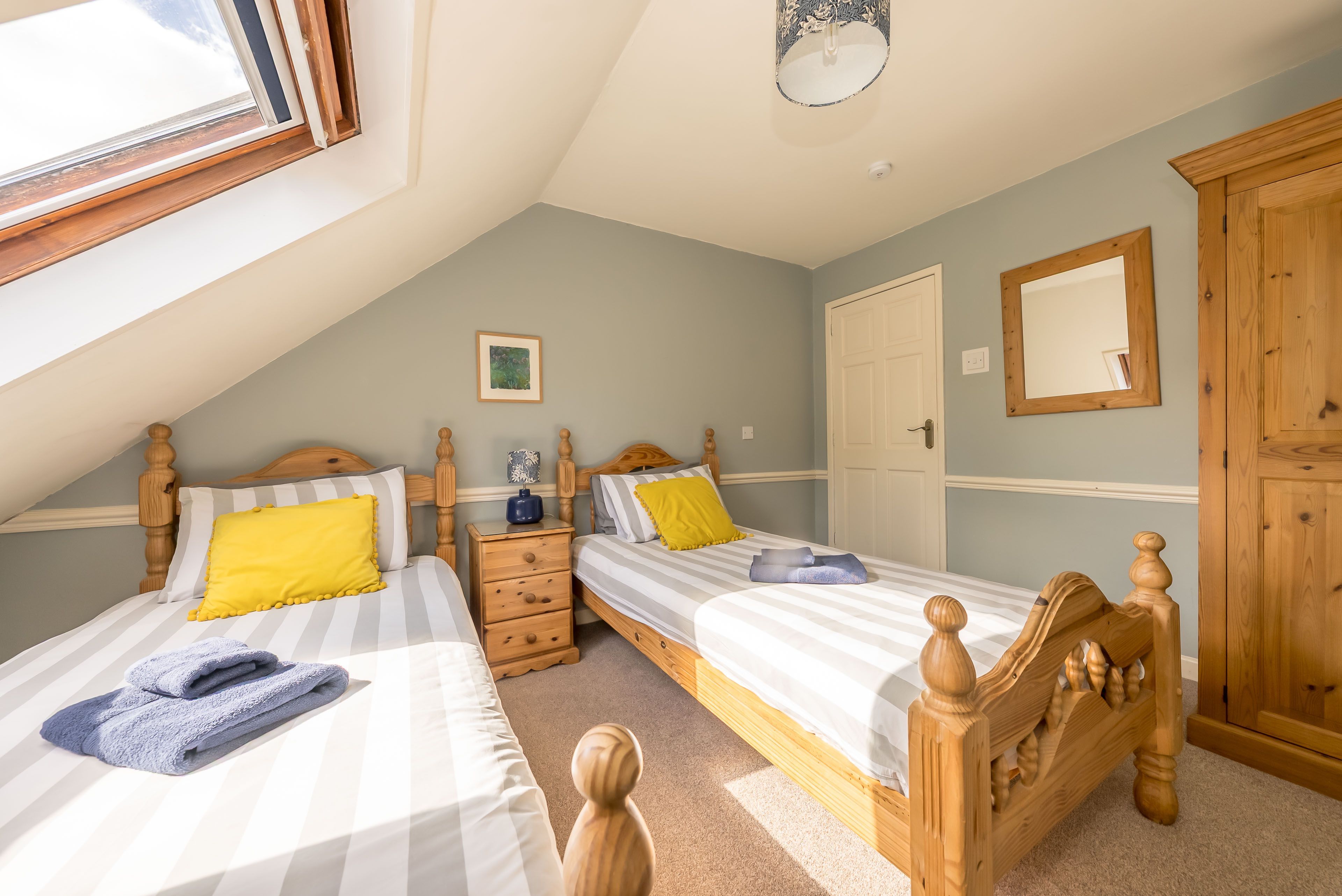 A bedroom with two beds, a wooden wardrobe, a bedside table, a framed picture, and a window.