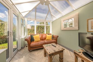 A conservatory with a glass ceiling, a brown leather sofa with cushions, a wooden coffee table, and a television on a wooden stand.