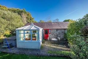 A small, enclosed garden area with a gazebo, a stone building with a red door, and a brick patio. - Romantic stone cottage with hot tub in Pembrokeshire | Eiddew-Y-Ffynnon (Pembrokeshire)
