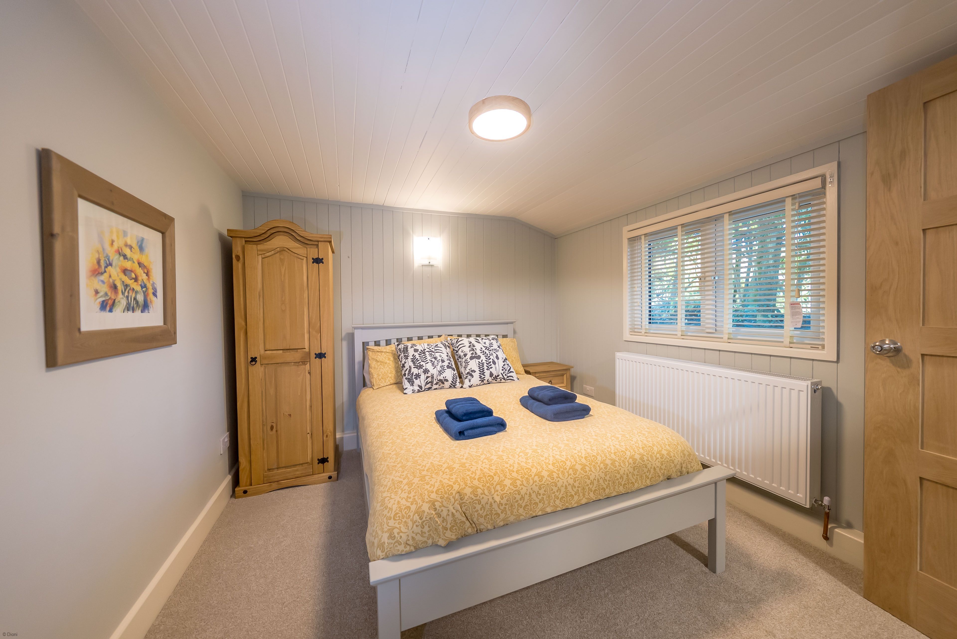 A bedroom with a bed, a nightstand, a wooden wardrobe, a framed picture, a window with blinds, and a ceiling light.