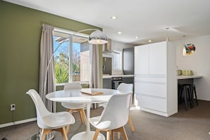 A modern kitchen with white cabinets, a dining area with a round table and chairs, and a view of the outdoors through large windows.