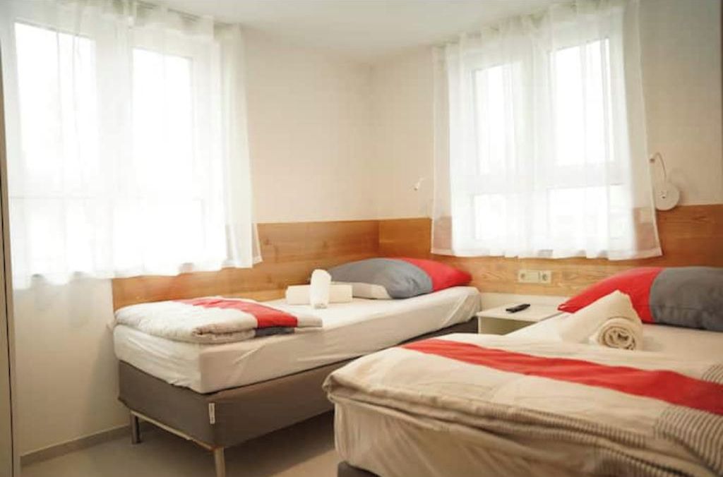 A room with two beds, each with a pillow and a blanket. There are curtains on the windows, and a lamp on the wall.
