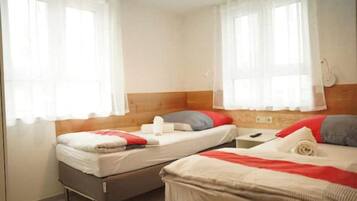Ein Zimmer mit zwei Betten, jeweils mit Kopfkissen und Decke. Vor den Fenstern hängen Vorhänge und an der Wand steht eine Lampe.