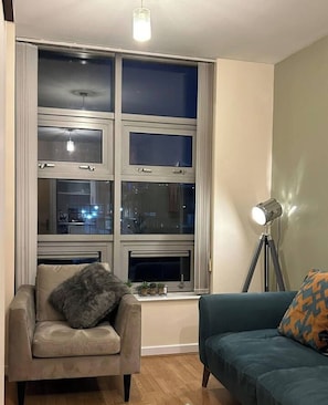 A room with a large window, a chair, a sofa, a floor lamp, and a small table with plants. - Central Birmingham Flat with Free Parking! (West Midlands)