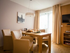 Un salon moderne avec une table à manger en bois, deux fauteuils beiges, un téléviseur à écran plat fixé sur un panneau en bois et une grande fenêtre ornée de rideaux légers.