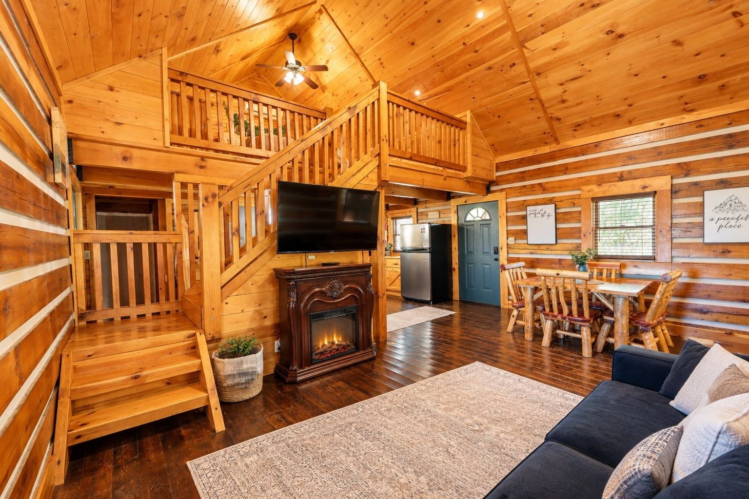 A wooden cabin interior with a fireplace, a TV, a dining area, and a staircase.