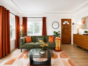 Ein Wohnzimmer mit einem grünen Sofa, orangefarbenen Kissen, einem Glastisch und einem gemusterten Teppich.