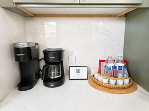 Eine Küchenzeile mit einer Keurig-Kaffeemaschine, einer Mr. Coffee-Kaffeemaschine, einem QR-Code für Hotelinformationen und einem Korb mit Wasserflaschen und Joghurtbechern.