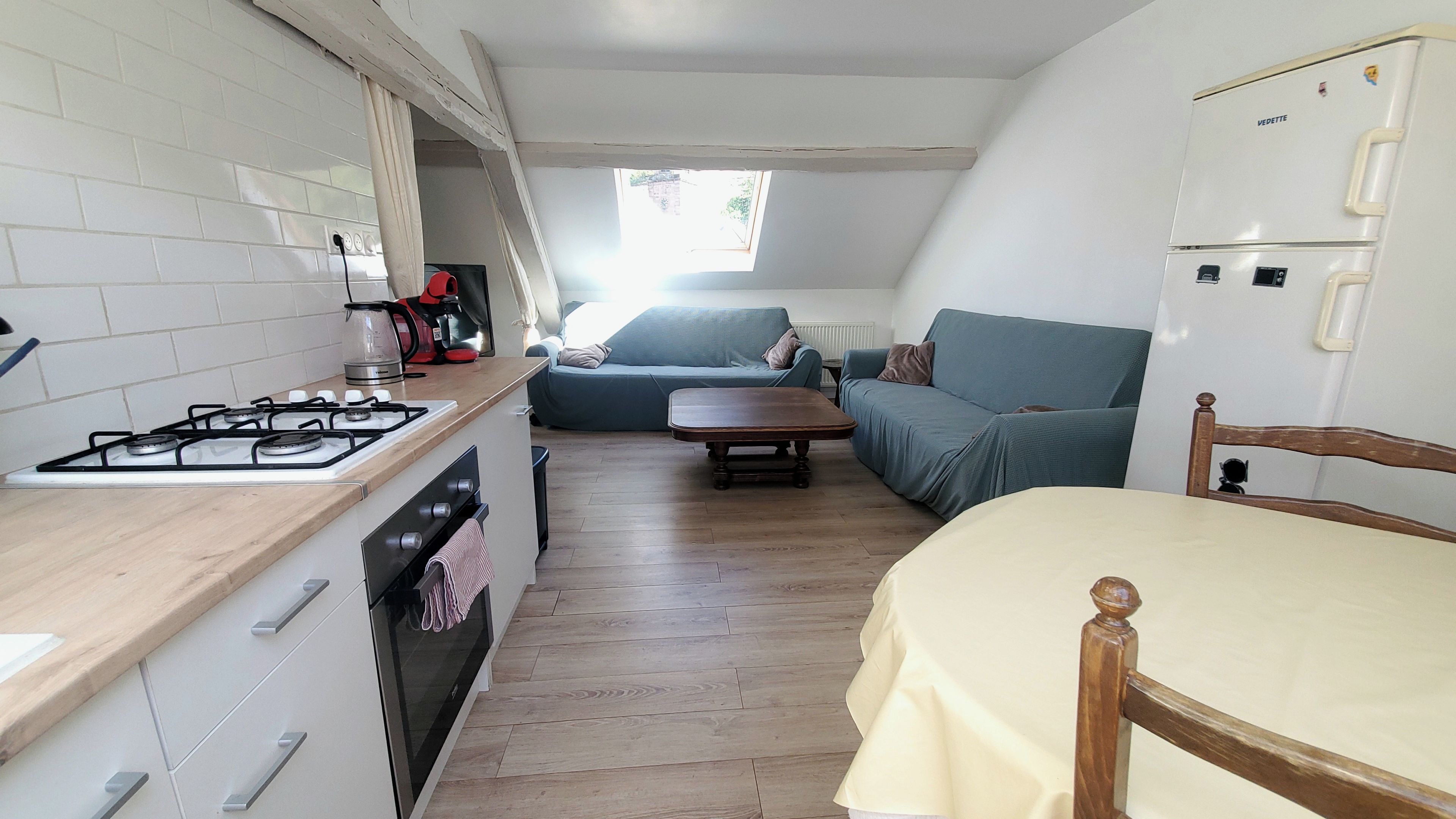 Cocina compacta con estufa a gas, horno y cafetera. Hay una zona de comedor con mesa y sillas, y un sofá al fondo.