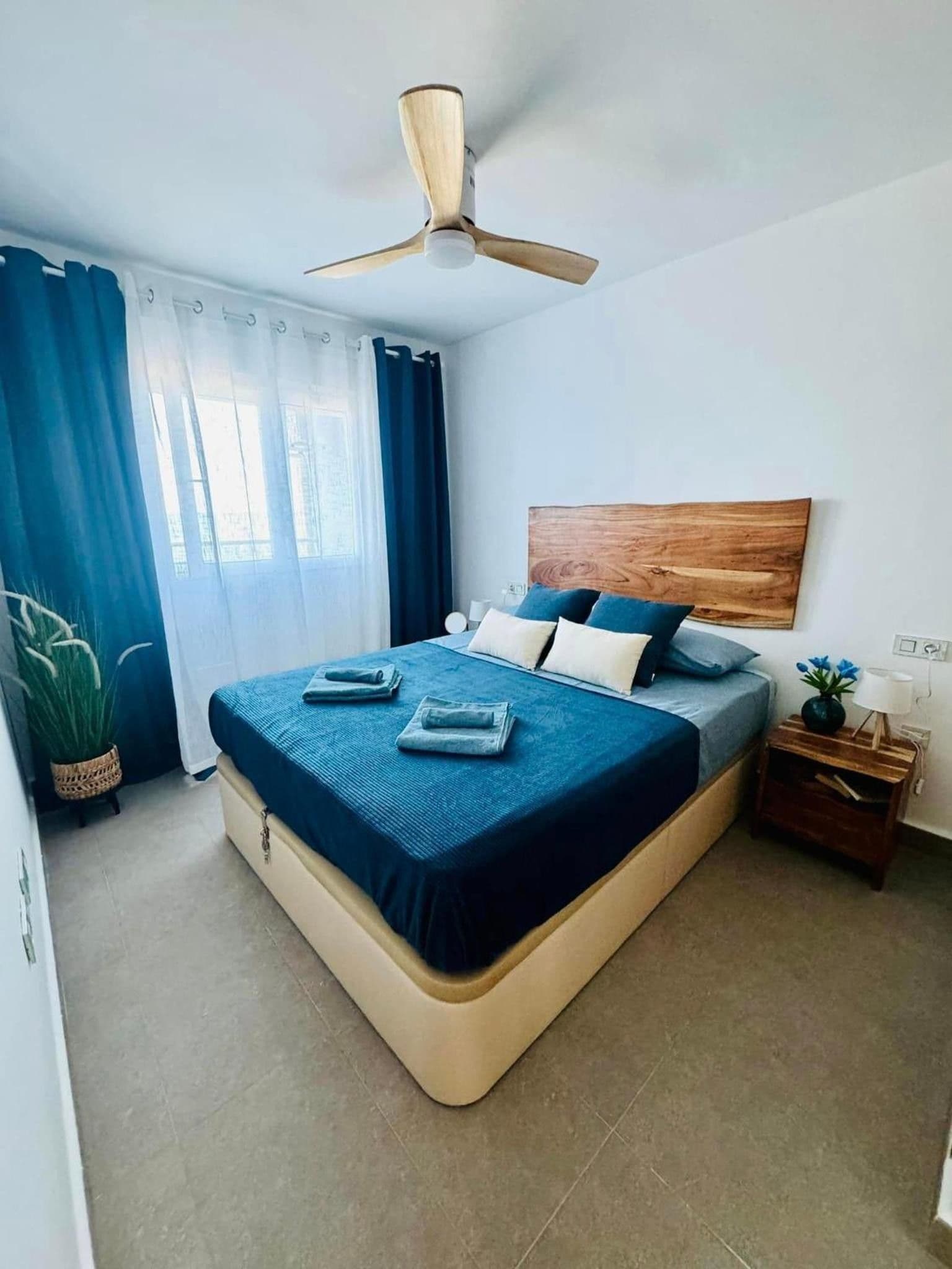 Ein Schlafzimmer mit einem Bett, einem Deckenventilator, einem Nachttisch, einer Topfpflanze und einem Fenster mit Vorhängen.
