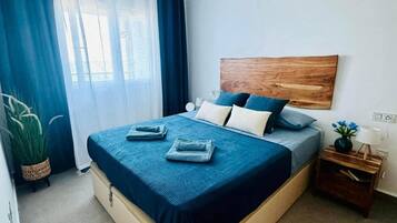 Ein Schlafzimmer mit einem Bett, einem Deckenventilator, einem Nachttisch, einer Topfpflanze und einem Fenster mit Vorhängen.