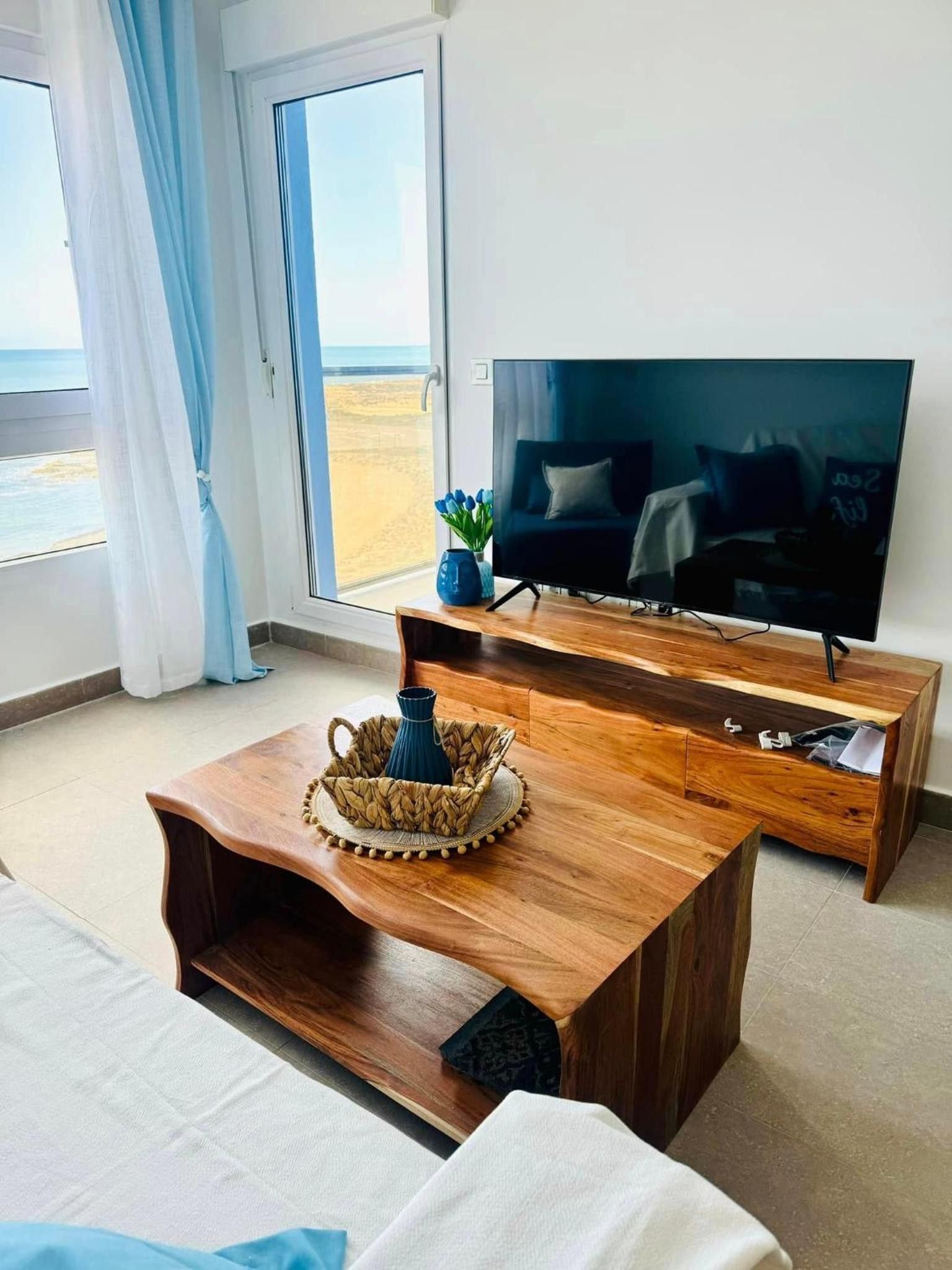Ein modernes Wohnzimmer mit einem großen Flachbildfernseher, Holzmöbeln und Blick auf den Strand durch eine Glastür.