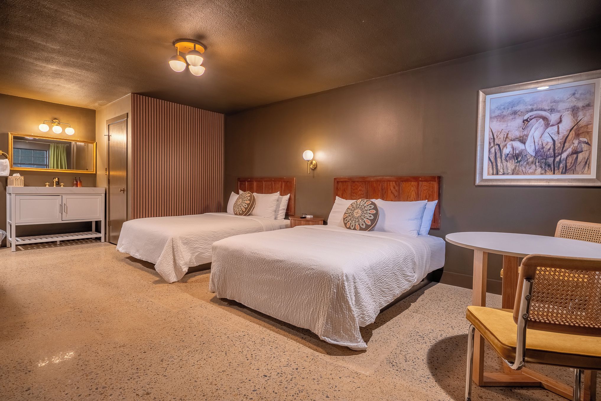 Quarto de hotel com duas camas, banheiro com toucador, uma mesa pequena e um quadro na parede.